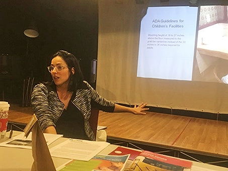 A woman with dark hair and glasses sits at a table delivering a workshop. She points to a screen that reads, "ADA Guidelines for Children's Facilities"